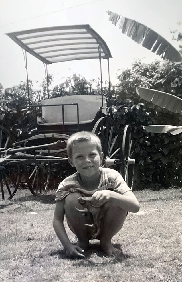 black and white photo of boy crouching outside next to a cart and large bushes. 