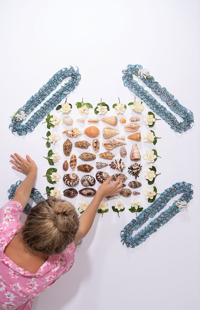 person in pink shirt arranges shells organized by colors of rainbow with blue leis arranged in symmetrical order. 
