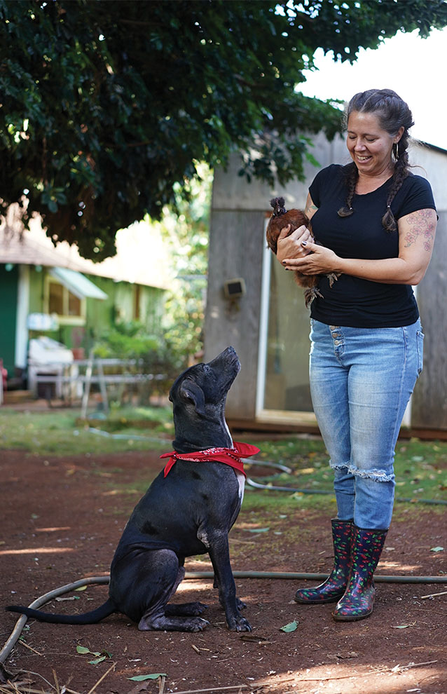 woman smiles and holds chicken while black dog sits looking up at them in the backyard. 