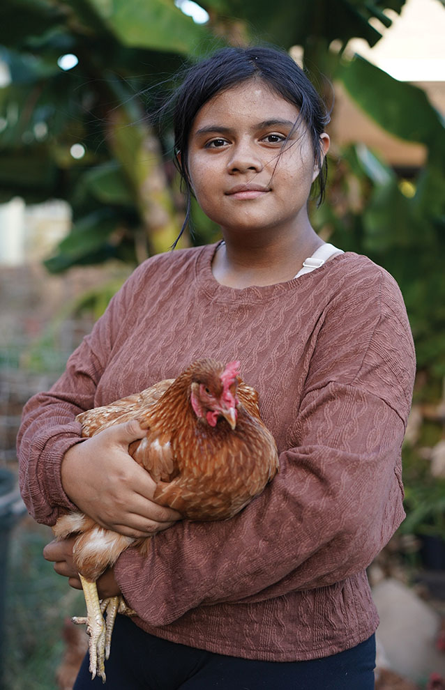 Young woman in a brown sweater holds a chicken and smiles gently against a tree in the background