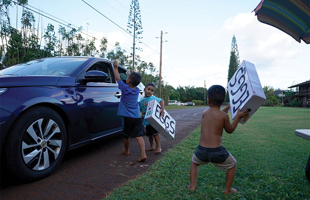 three kids hold eggs for sale sign as a car drives past them on a country road. 