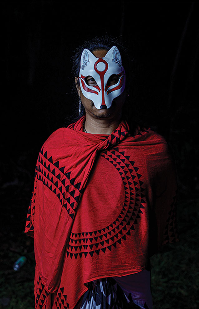 traditional red shawl is draped over a man with a mask against a black background. 