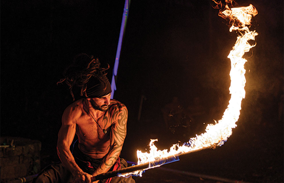 a tattooed fire artist holds fire stick as fire trails out behind him. 