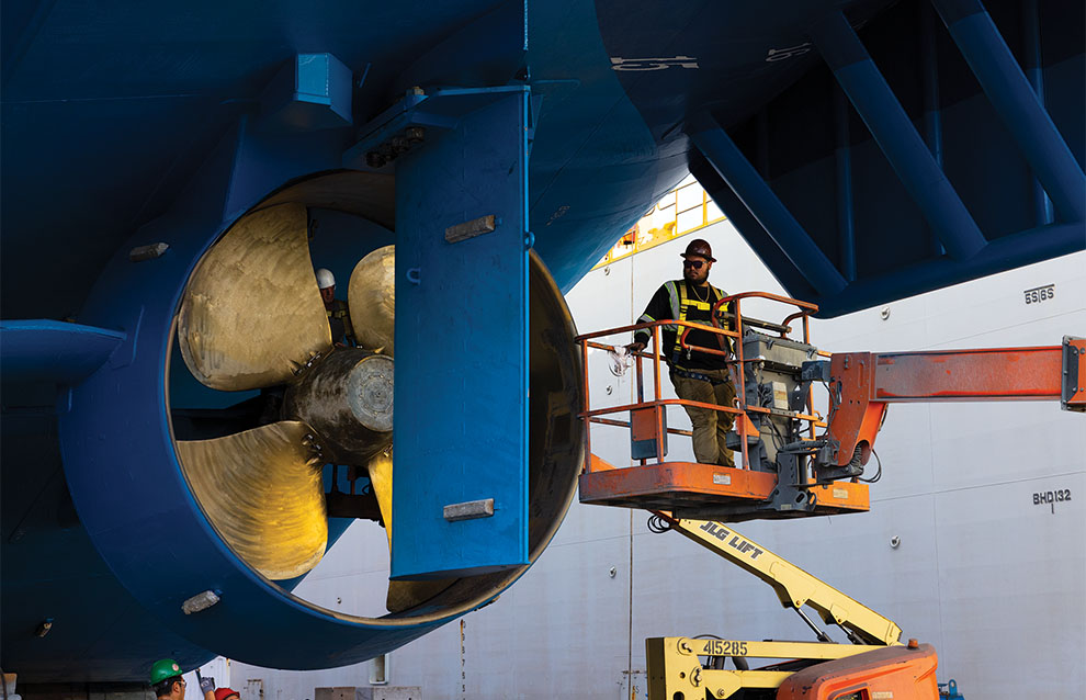 a construction worker stands next to the blue and gold propeller on the underside of the boat. 