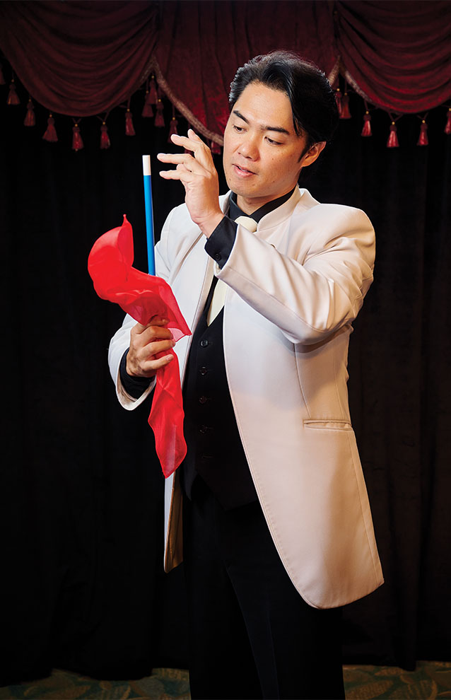 magician in white jacket holds magician's wand with a red tail of feathers attached. 