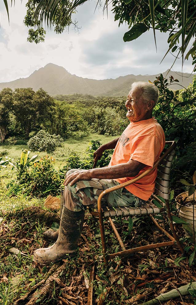 older man in bright orange t-shirt sits outside on a wooden chair with the Hawaiian mountains and lush landscape in the background. 